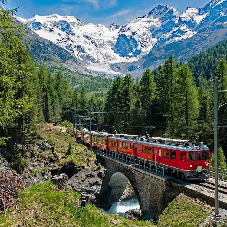 Red train crossing a stone bridge below snowy mountains and a glacier